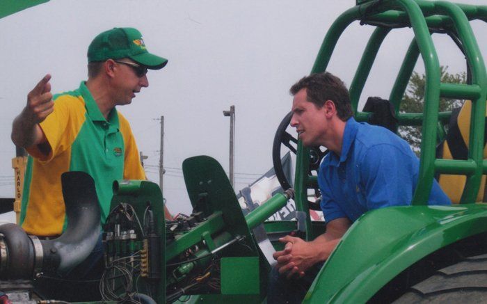 Two men by a green tractor; one gestures, the other listens. Outdoors on a cloudy day.