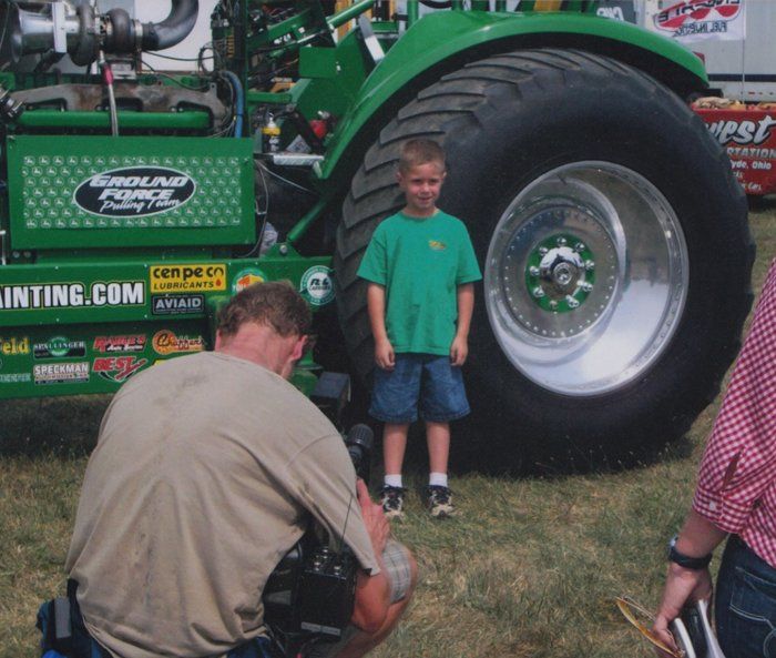 Boy stands in front of large tractor tire; man crouches taking a photo. Green tractor, sunny outdoors.