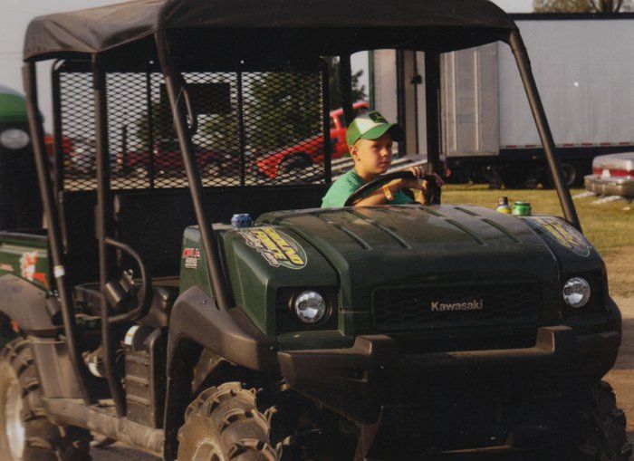 Boy driving a green Kawasaki utility vehicle outdoors, wearing a hat, smiling.
