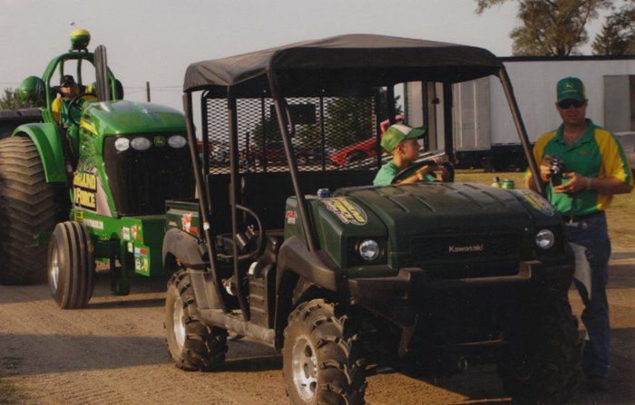 A green utility vehicle with a person driving, and a tractor in the background. Man standing by the vehicle.