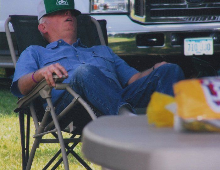 Man asleep in a folding chair, wearing a blue shirt and jeans, resting outside.