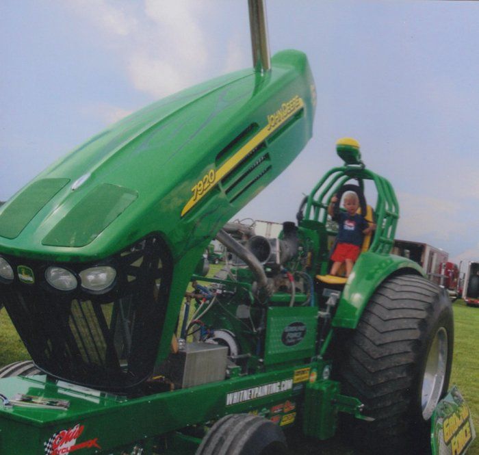 Green John Deere tractor with open hood, child in seat. Outdoor setting.