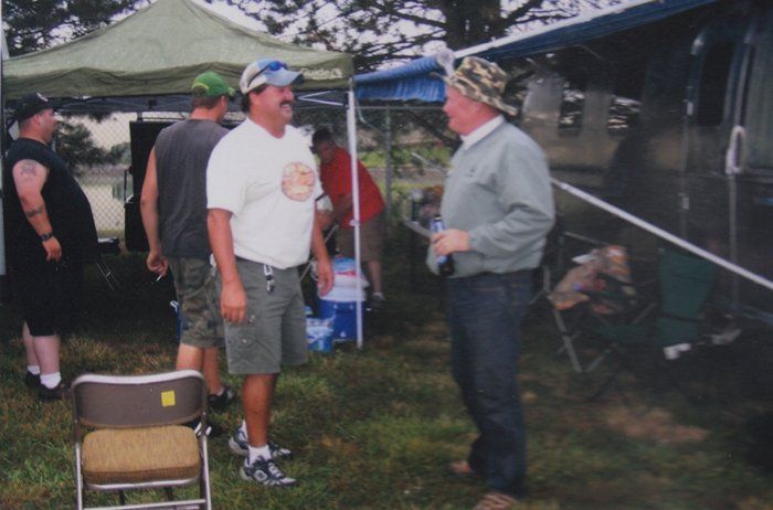 People talking outside near tents and a vehicle on a grassy area.