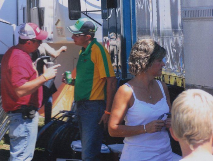 People socializing outdoors near a vehicle. One man in pink shirt and another in green/yellow shirt are talking. 