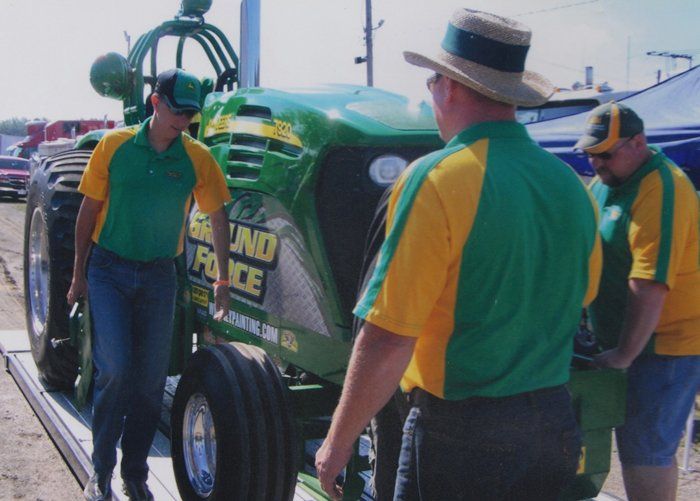 Three men by a green and yellow tractor; one wearing a straw hat. They are at an outdoor event.