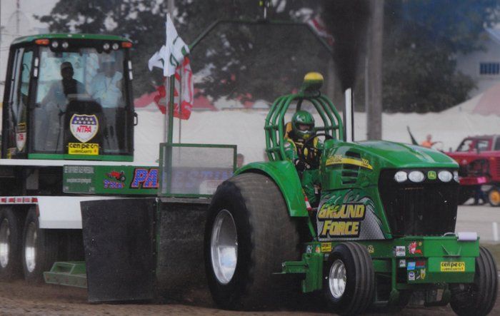 Green tractor pulling a weight sled, billowing black smoke, at a tractor pull event.