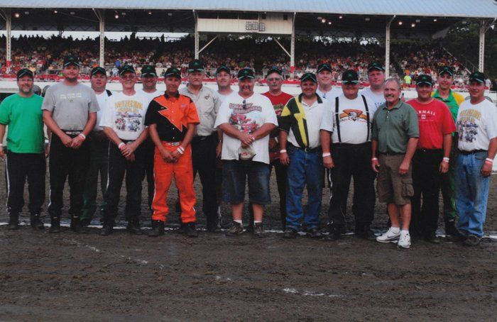 Group of men posing on a dirt track, some wearing hats and t-shirts, stands in front of a grandstand filled with people.
