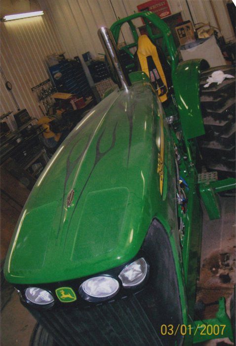 Green John Deere tractor with custom flames and a chrome exhaust stack, in a workshop.