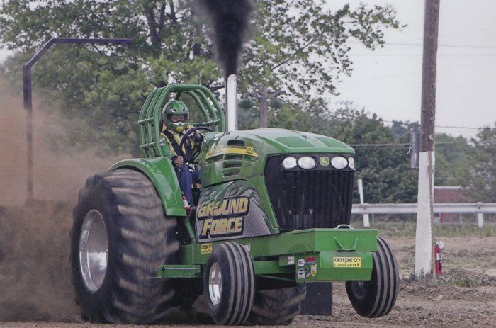 Green tractor pulling, belching black smoke. Driver in helmet. Ground Force logo on tractor.