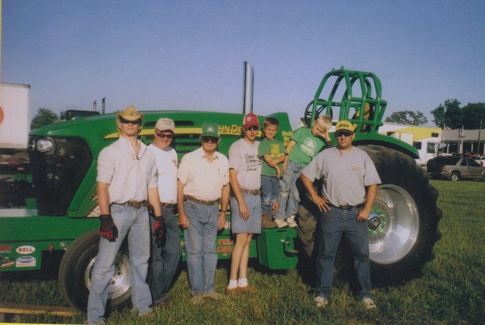 Group of people posing with a large green tractor at an outdoor event.