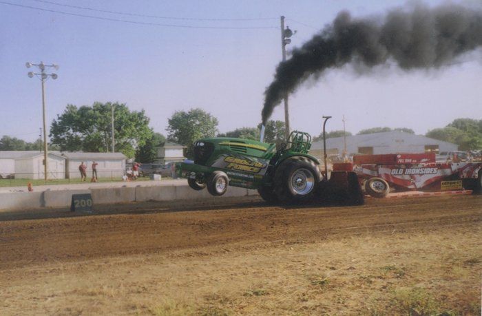 Green tractor with black smoke pulling a sled in a dirt arena, front wheels raised.
