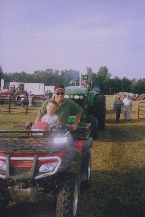 Man and child on a red ATV, green tractor in the background, fairground setting.