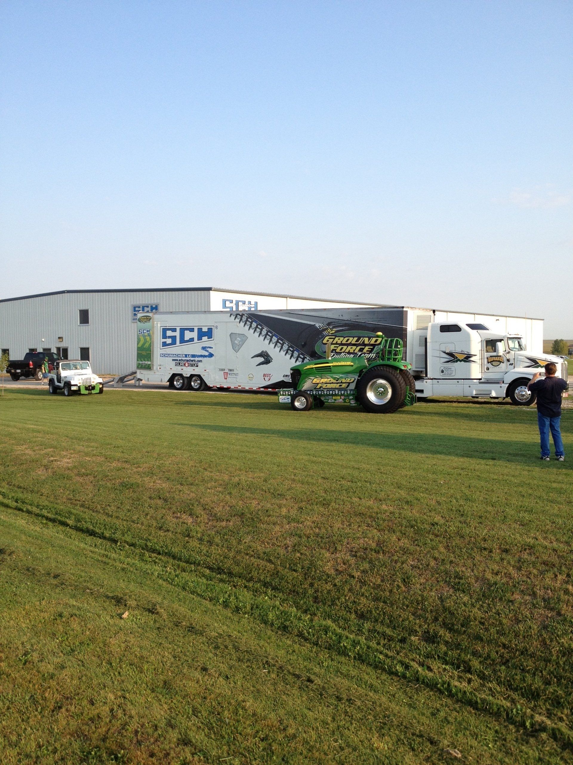 A tractor-pulling rig parked on grass. The trailer has a GCH logo and the tractor is green. A man stands nearby.