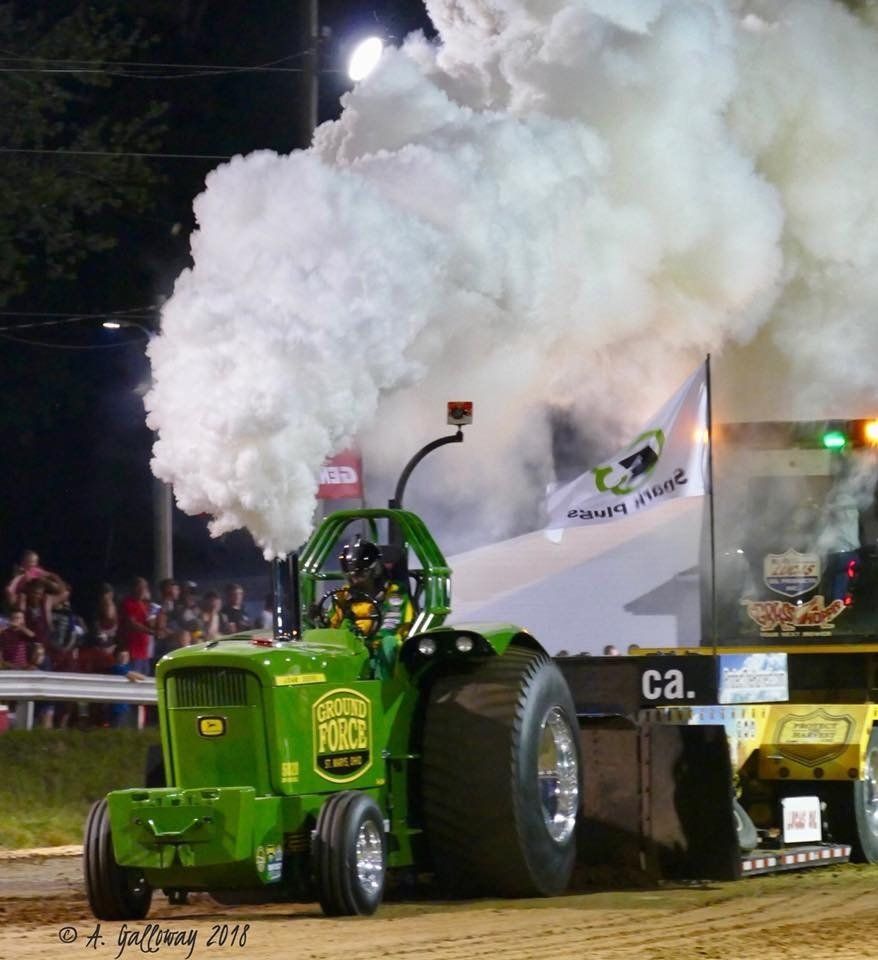 Green tractor pulling a heavy sled, billowing white smoke, at a nighttime tractor pull competition.