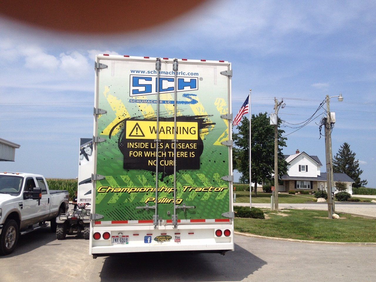 Rear of a white SCH Equipment semi-truck with warning sign, parked near a house, flag, and utility poles.