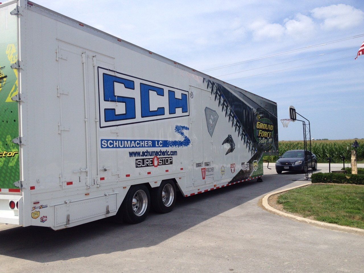 White Schumacher racing trailer with blue logo, parked outdoors. A car is nearby.