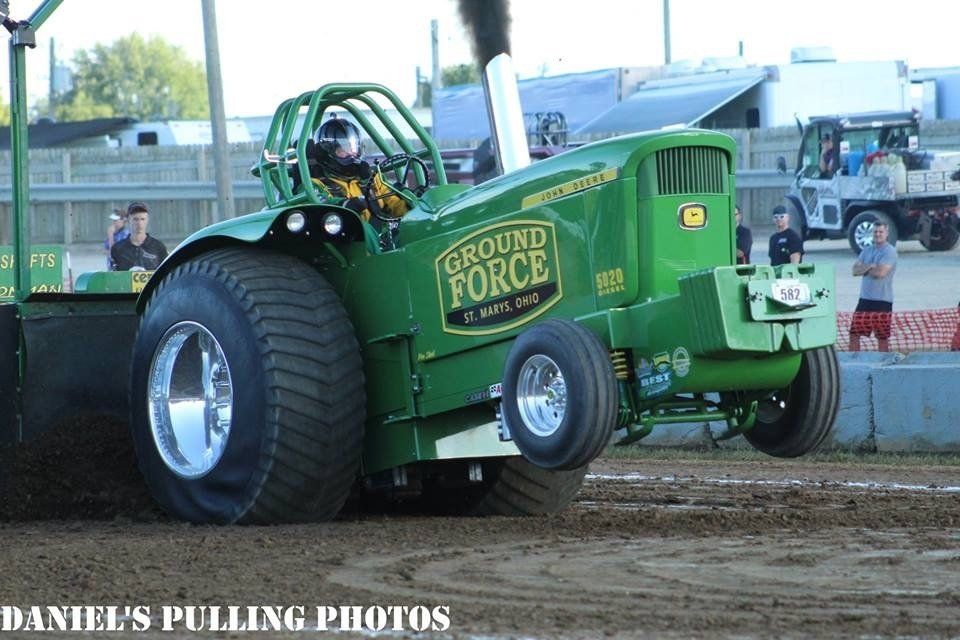 Green tractor doing a pull, black smoke billowing, driver visible. 