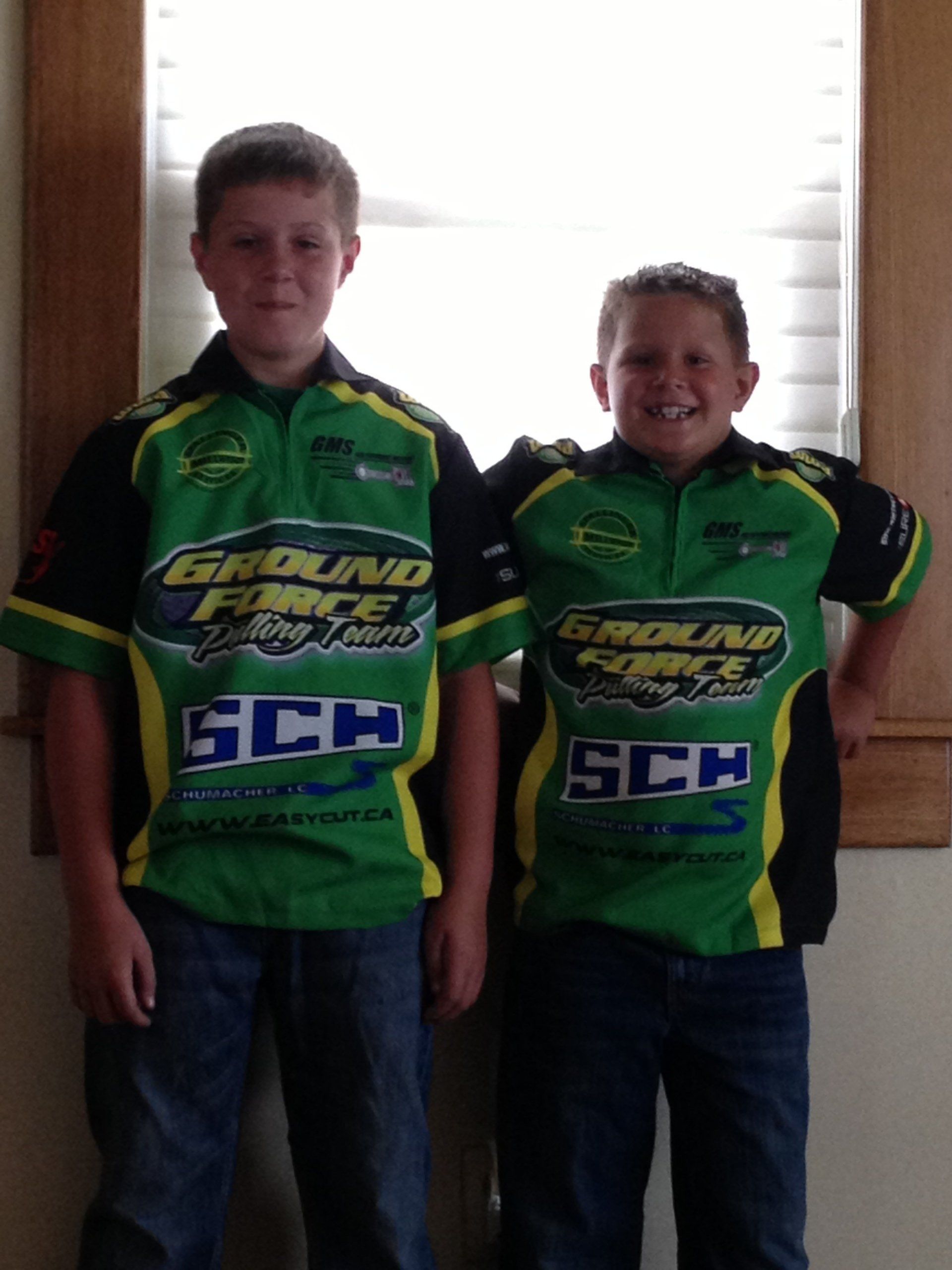 Two boys in matching green Ground Force shirts and jeans pose by a window.
