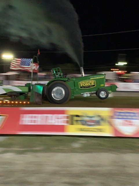 Green tractor in a pulling competition, emitting black smoke. American flag in background.