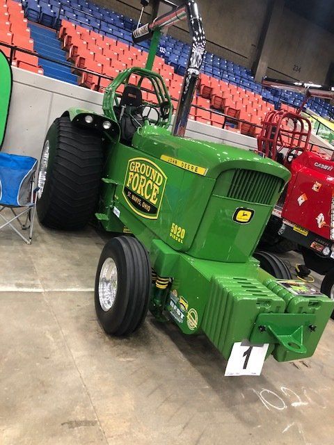 Green Ground Force tractor with large tires, indoors in front of stadium seating.
