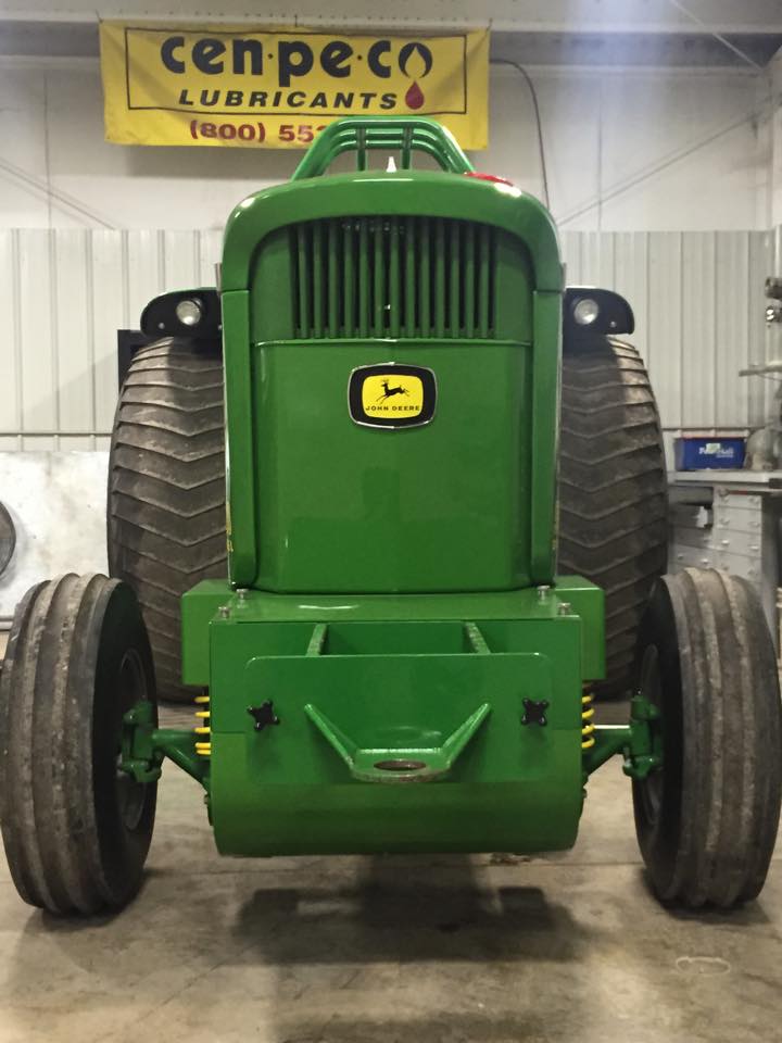 Green John Deere tractor, front view, in a workshop, tires, yellow sign in background.