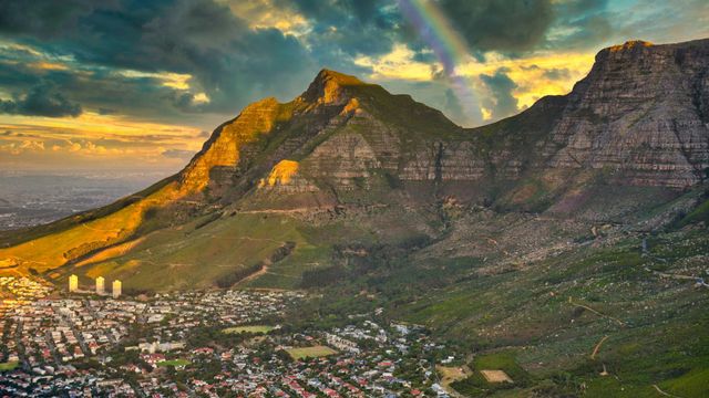 A mountain landscape at sunset with a faint rainbow in the sky, overlooking a residential neighborhood below.