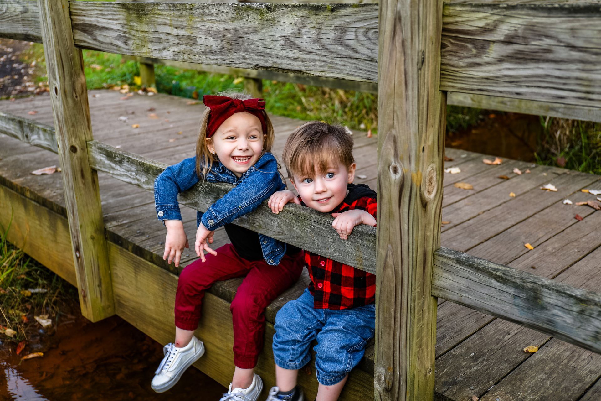 A boy and a girl are sitting on a wooden bridge over a river.