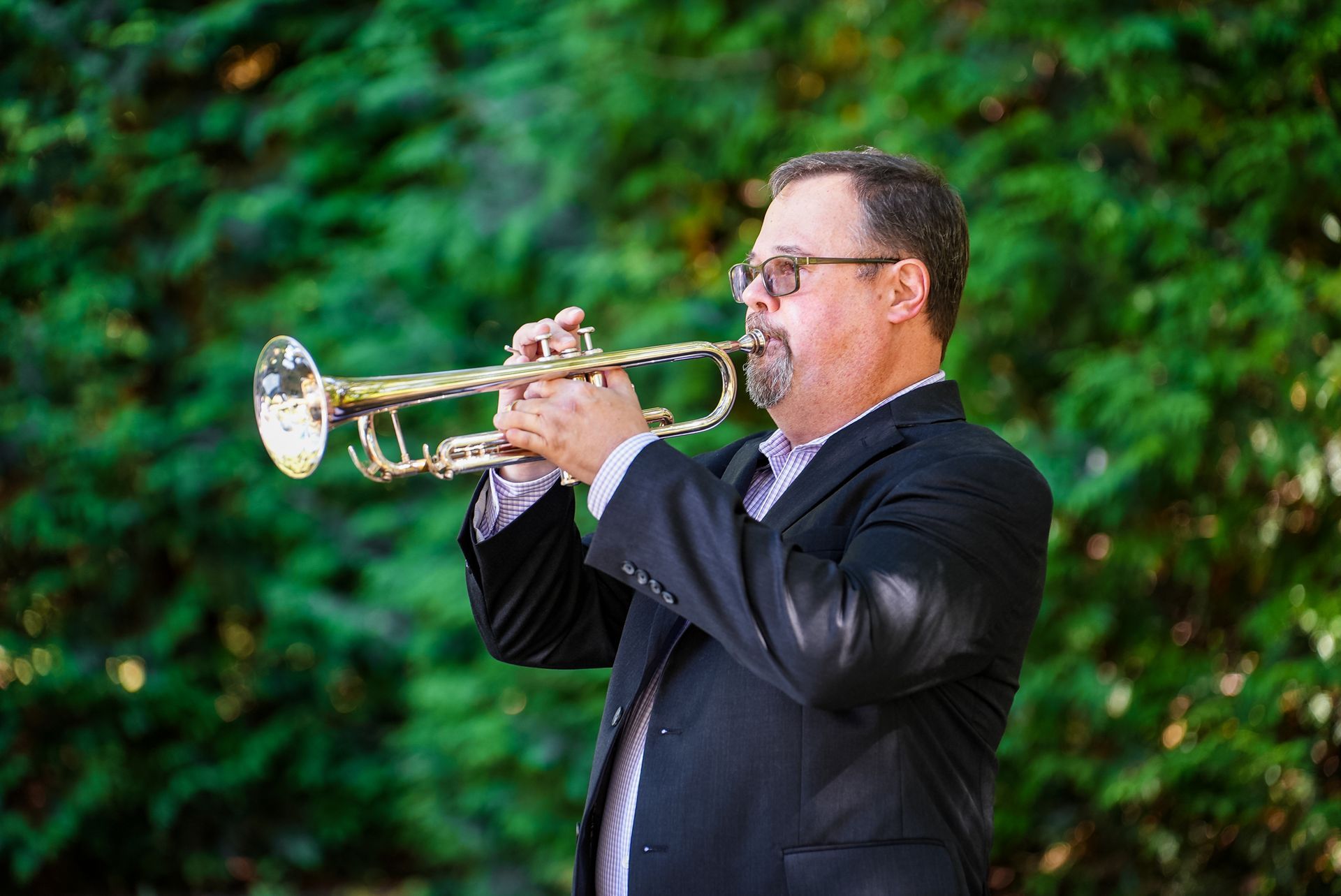 A man in a suit is playing a trumpet in a park.