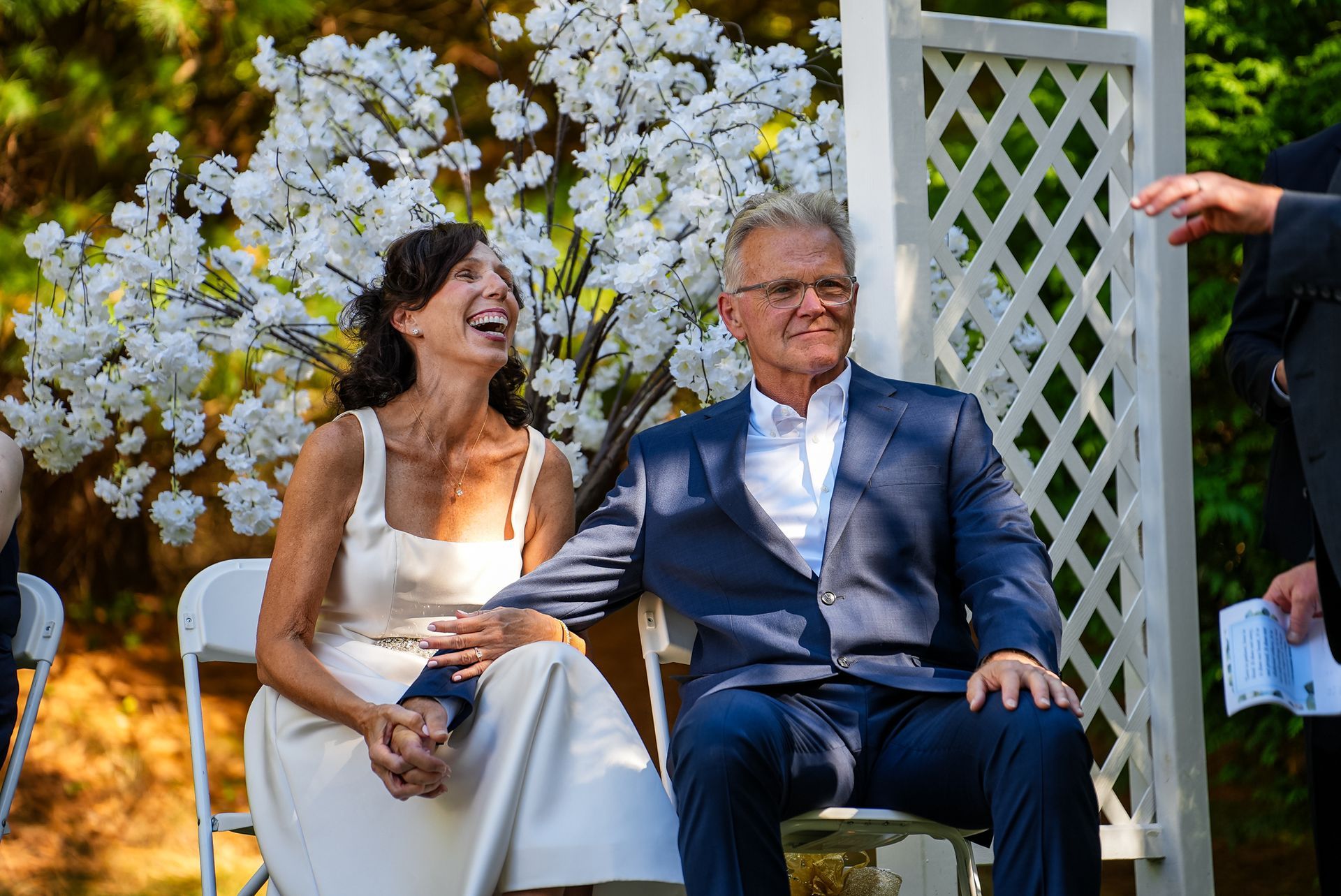 A bride and groom are sitting in chairs holding hands and laughing at their wedding ceremony.