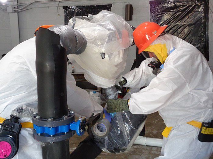 Two workers in white protective suits and hard hats inspect industrial piping in a room with black plastic-covered equipment.