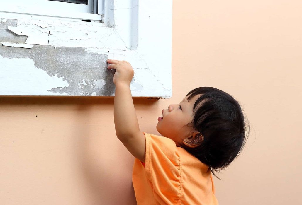 Child in an orange shirt reaching up to touch a peeling white window frame on a peach wall