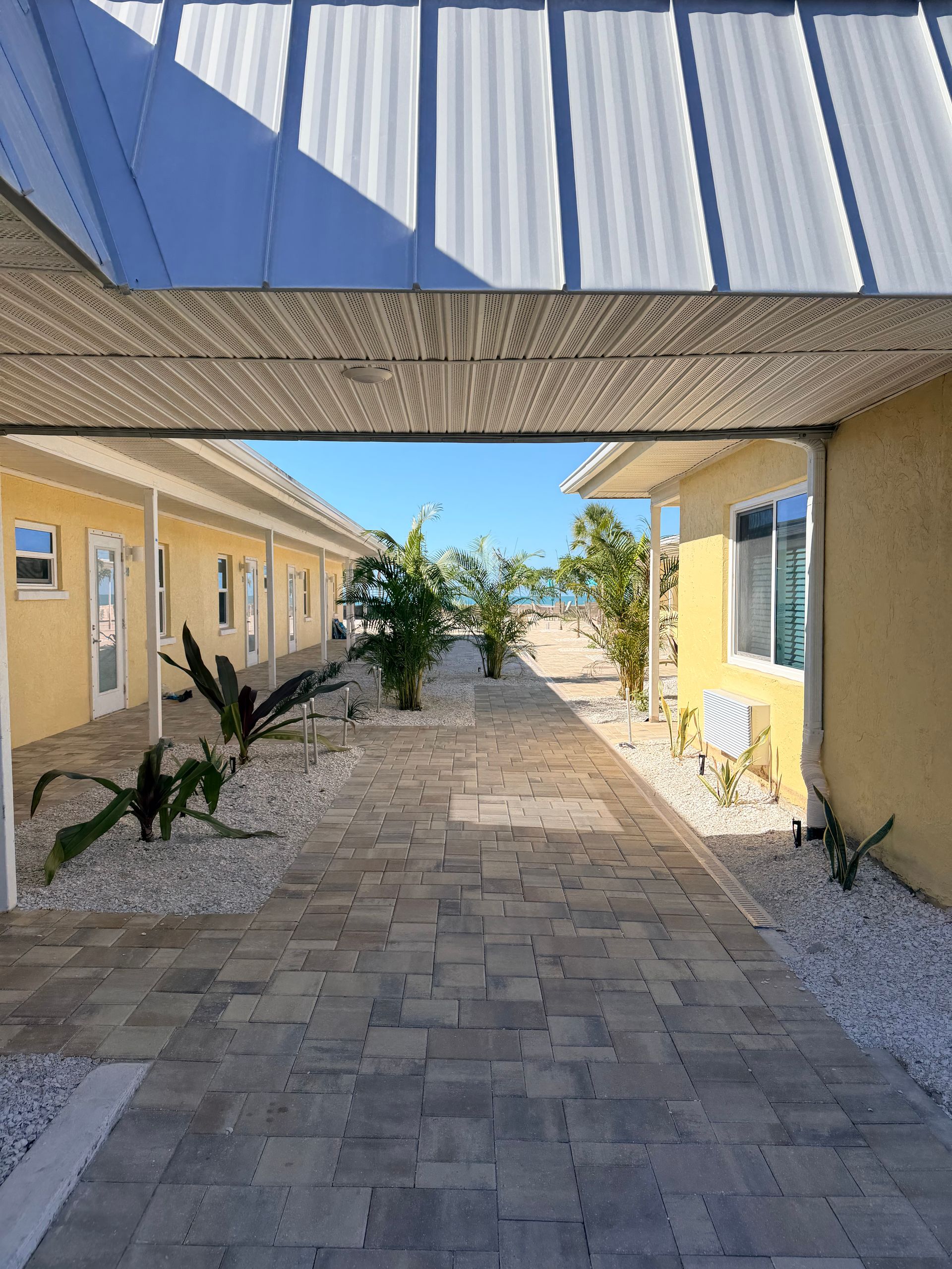 Brick path between yellow buildings, leading to a blue sky and water view, under a metal roof.
