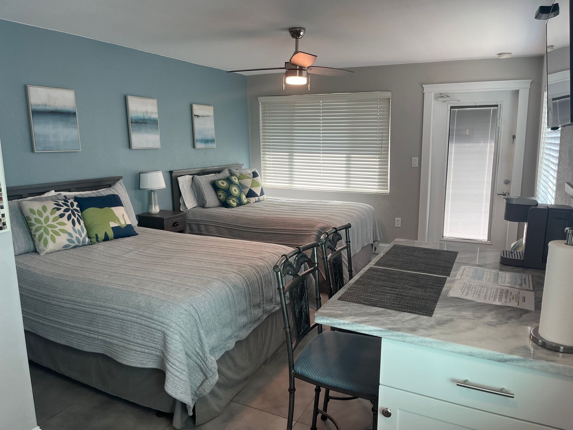 Studio bedroom with two beds, small kitchen area, blue walls, artwork, and a ceiling fan.