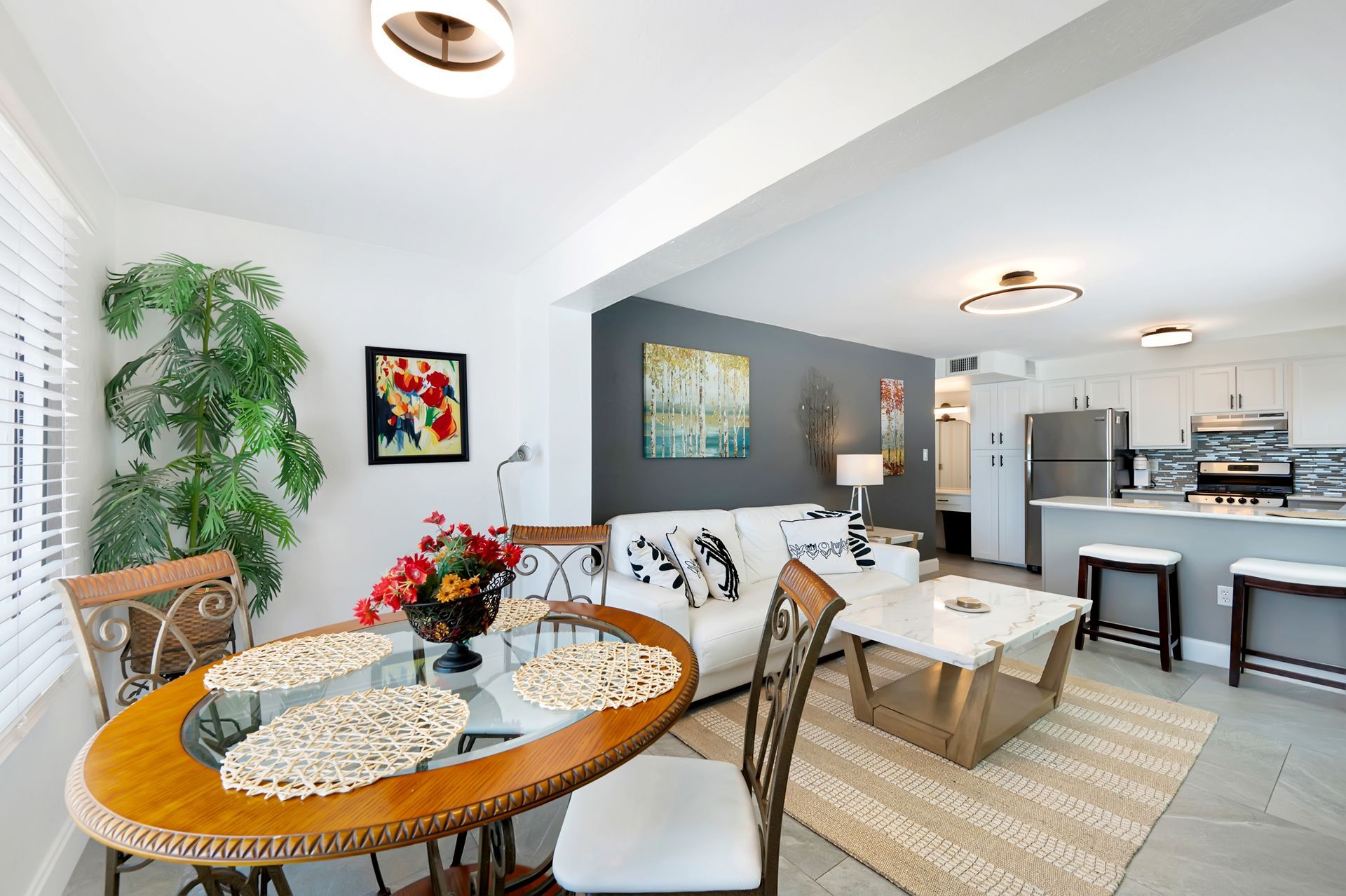 Open-concept living space with a dining table, sofa, and kitchen with a gray accent wall and natural light.