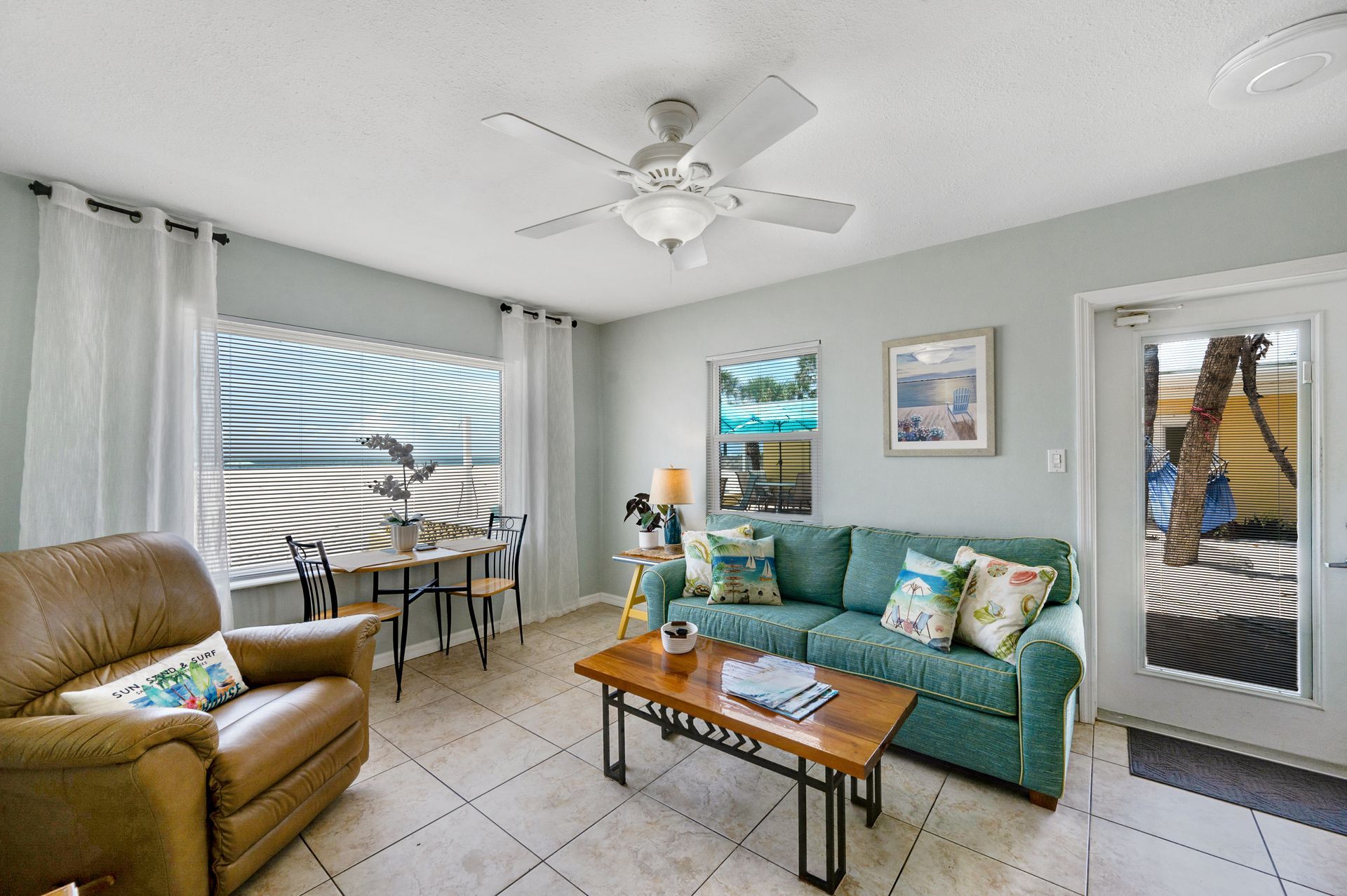 A bright living room with a leather recliner, teal sofa, coffee table, and a small dining area by the window.