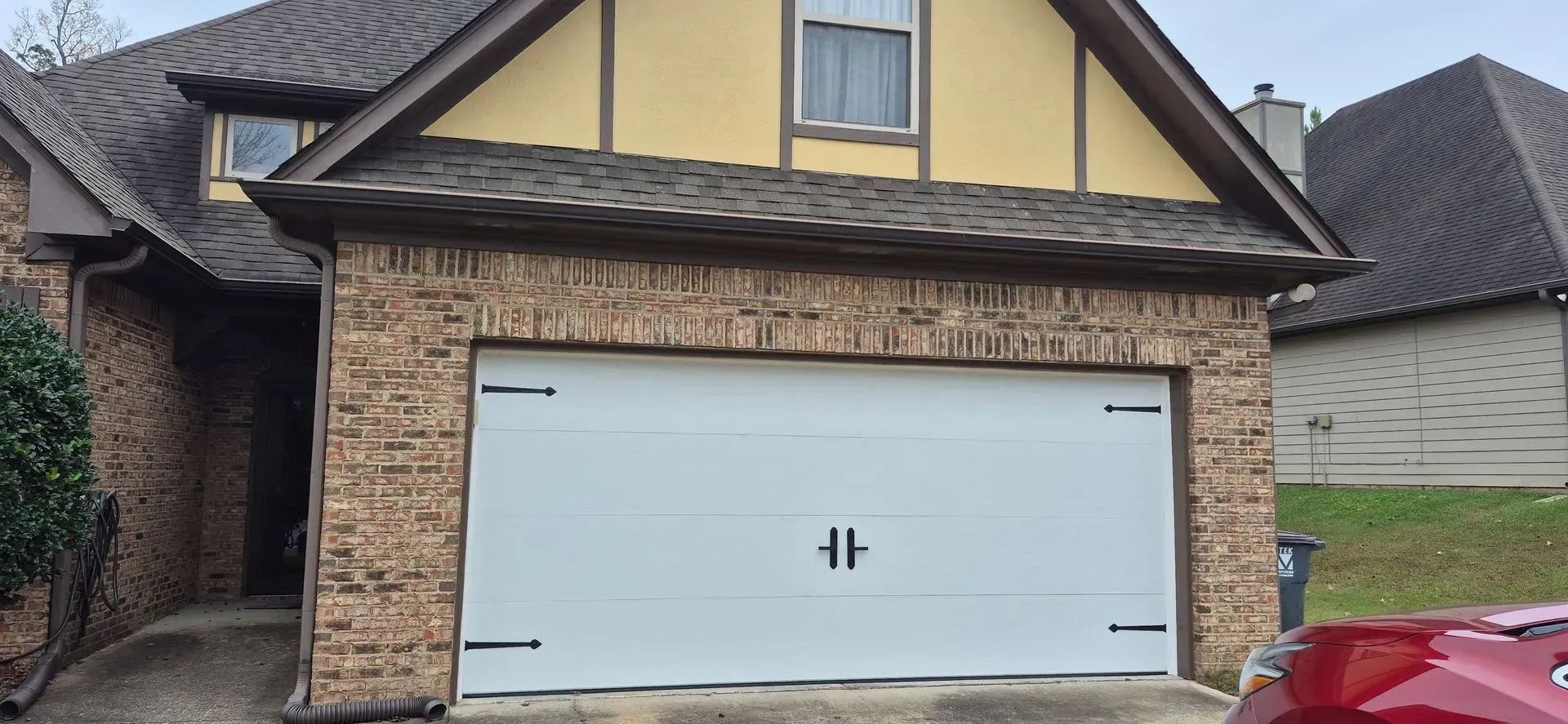 A red car is parked in front of a brick house with a white garage door.