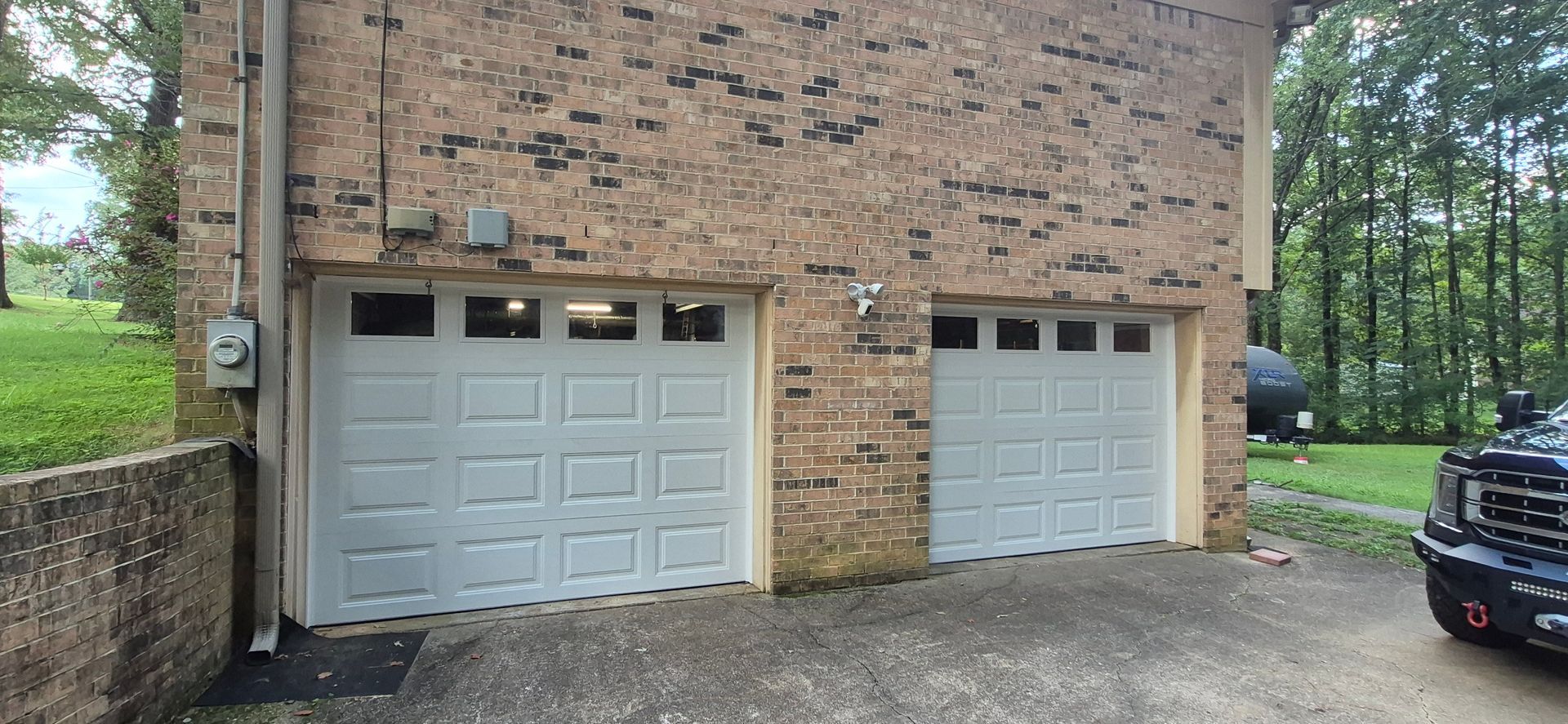 Two light gray garage doors on a brick building. A utility pole and a car are partially visible on either side.