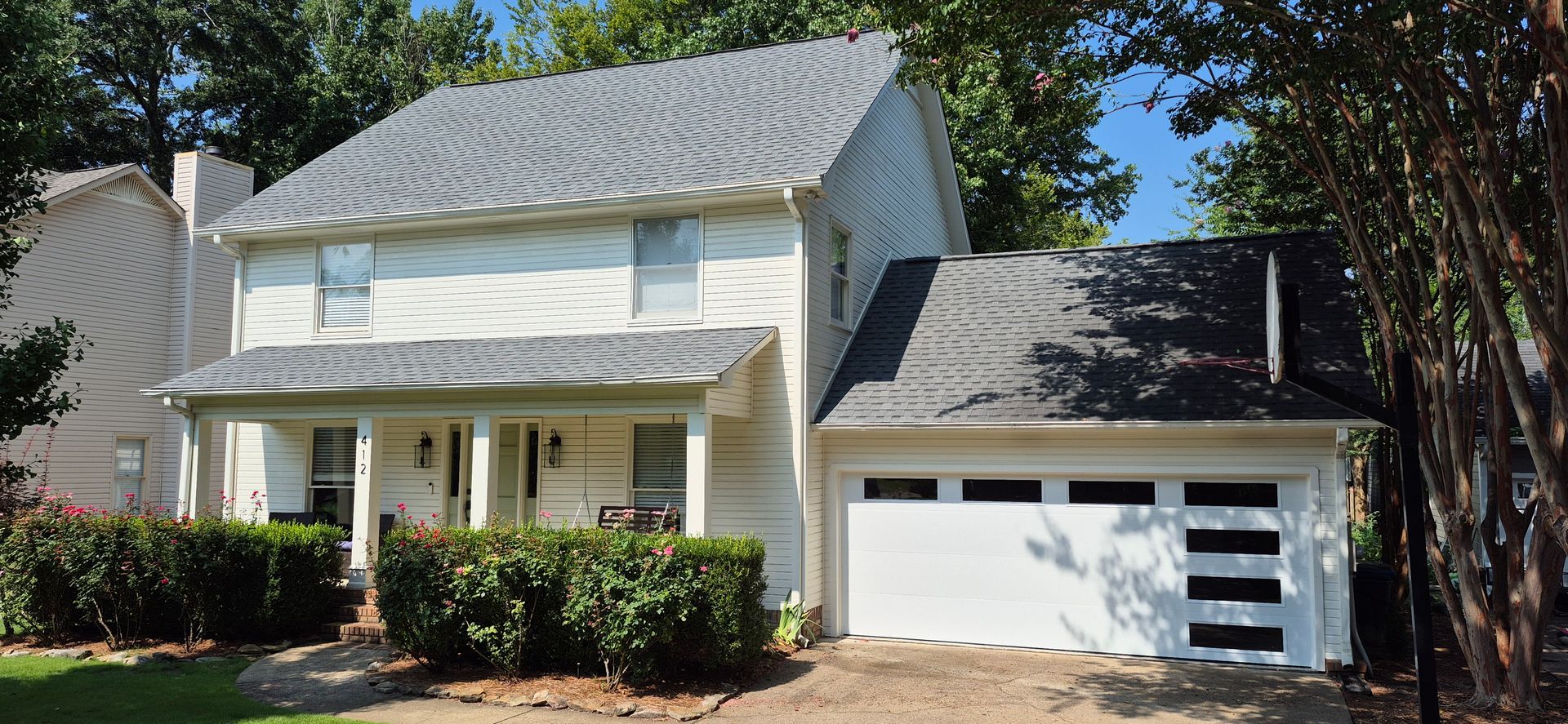 Two-story house with a white exterior, gray roof, and modern garage door. Lush green bushes line the front.