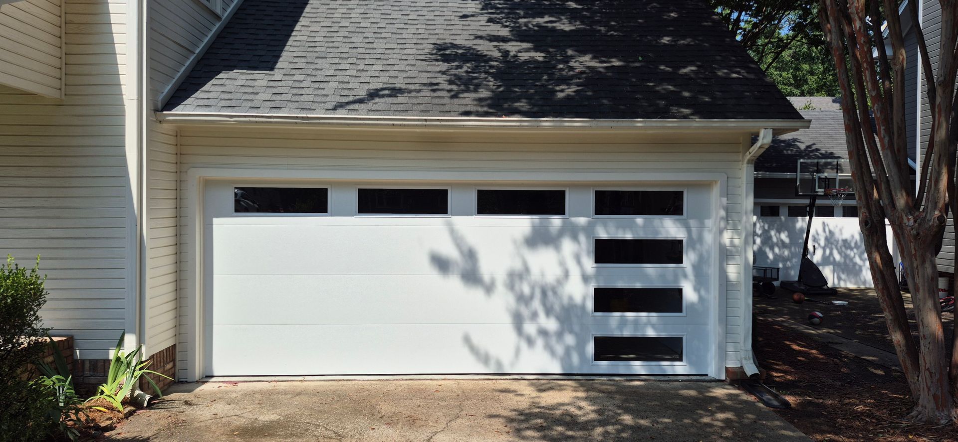 White garage door with black rectangular windows and a dark roof, in front of a gravel driveway, and trees casting shadows.