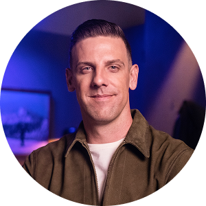 Smiling man in a dark jacket and black shirt, in front of a wooden wall and abstract art.
