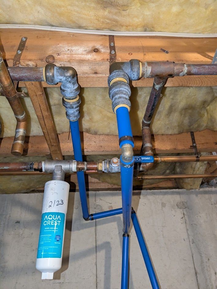 Under-sink plumbing with blue water filter, copper pipes, and shutoff valves against exposed wooden joists