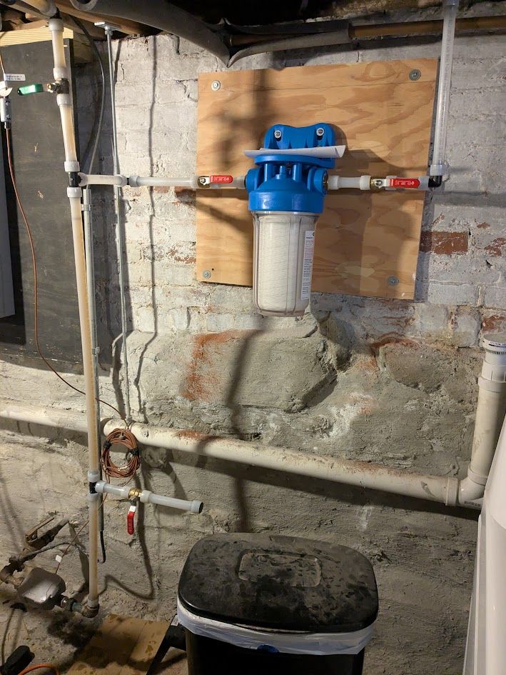 Basement utility wall with blue water filter and pipes above a black bucket on the floor