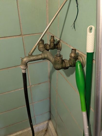 Old metal shower faucet with green-handled cleaning tools in a tiled bathroom corner