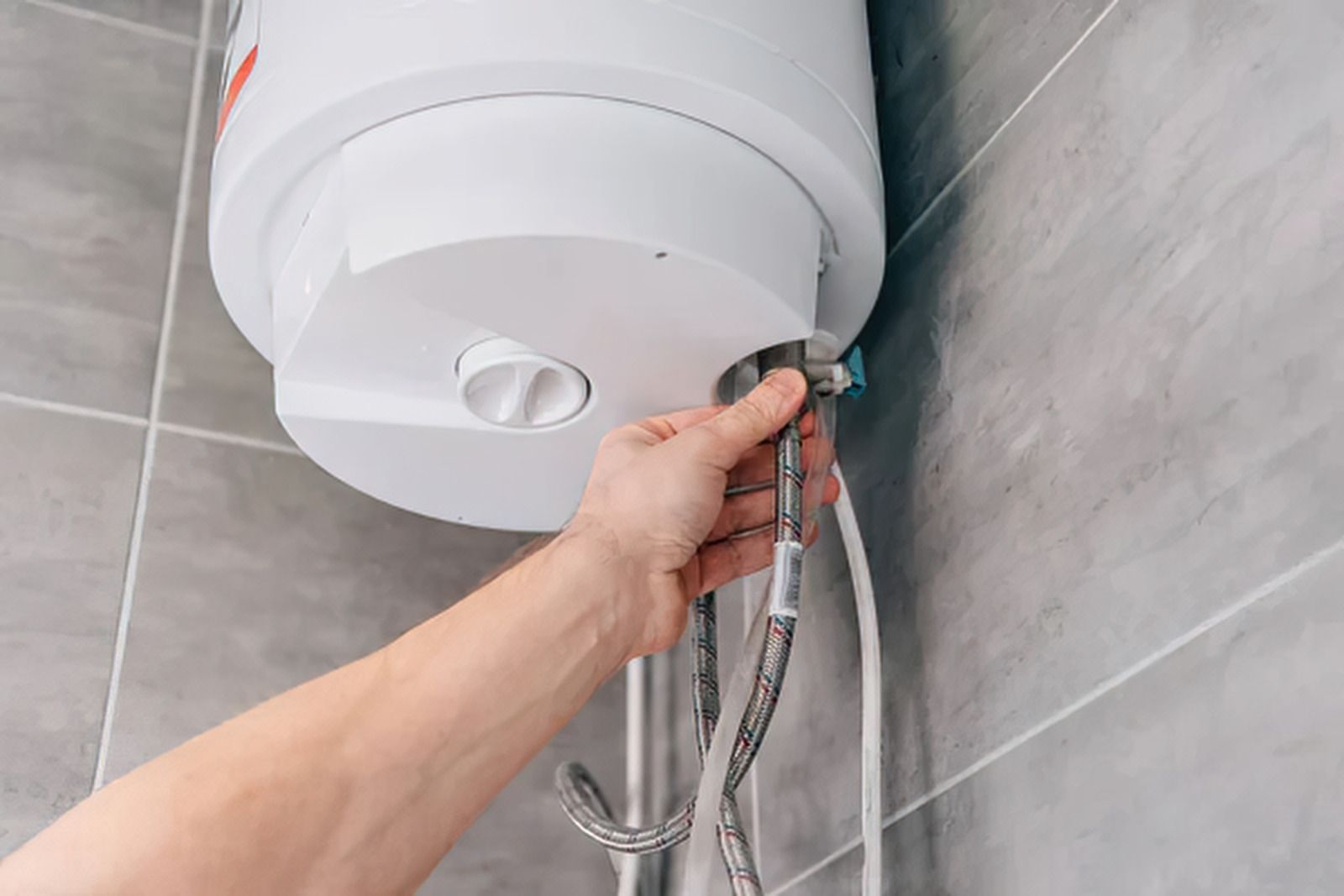 Hand adjusting a shower hose beneath a white bathroom water heater against gray tiled walls