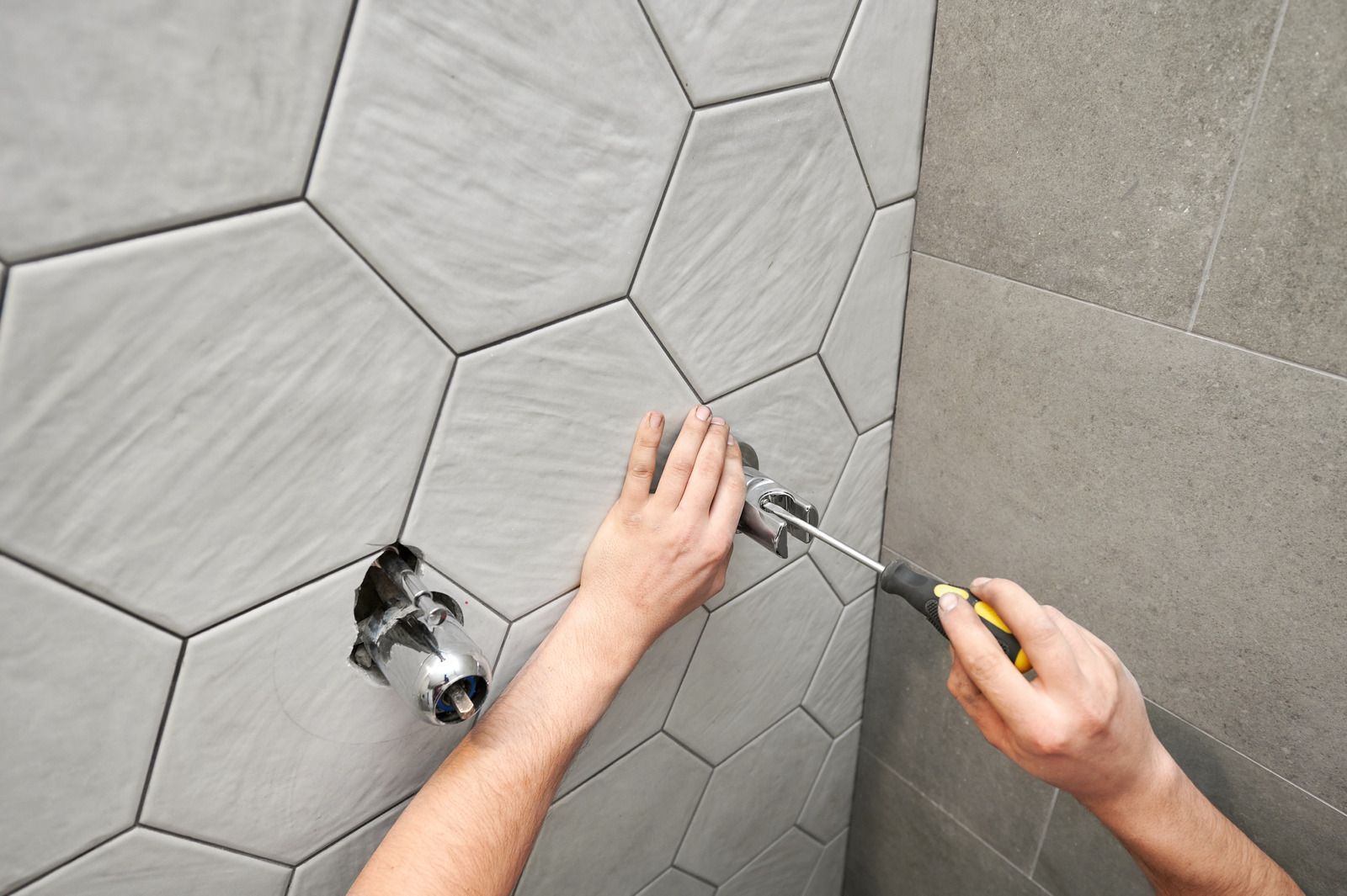 Hands installing hexagonal gray wall tiles with a trowel and utility knife.