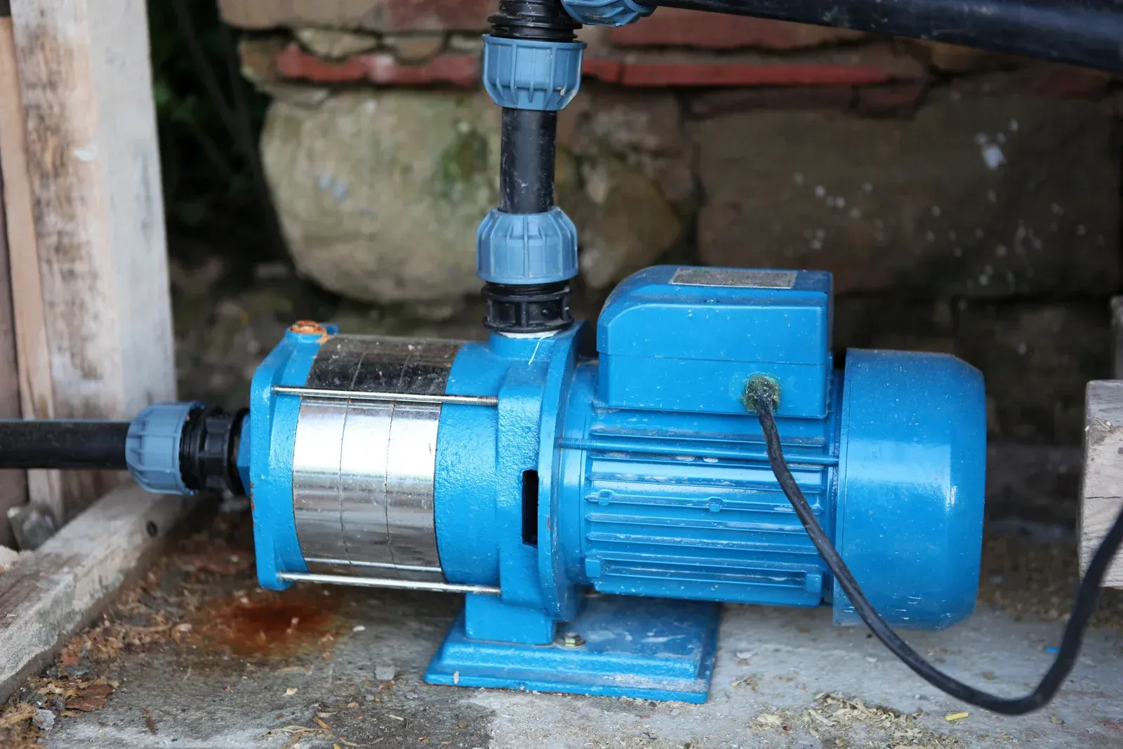 Blue electric water pump connected to pipes beside a brick wall on a concrete floor
