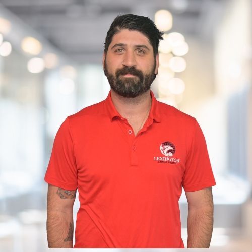 Man with beard in red polo shirt, arms crossed, in a blurred indoor setting.