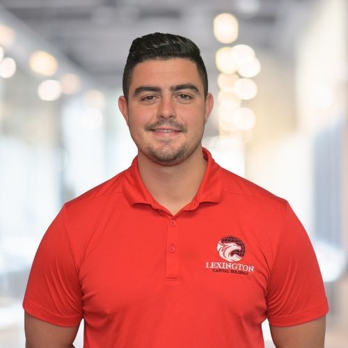 Man wearing a red polo shirt with a logo, smiling in a blurred office setting.