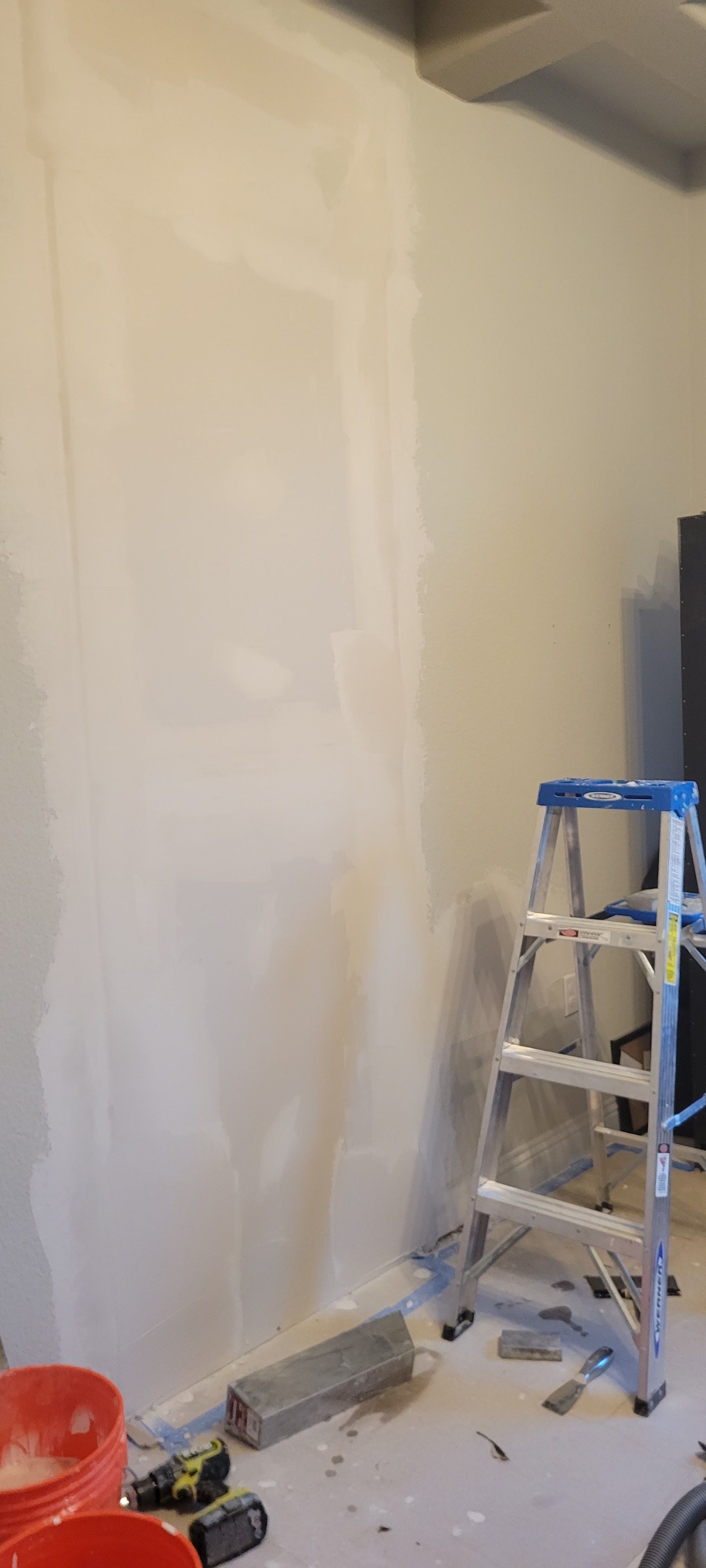 A room with drywall work in progress. A ladder, buckets, and tools sit on the floor.