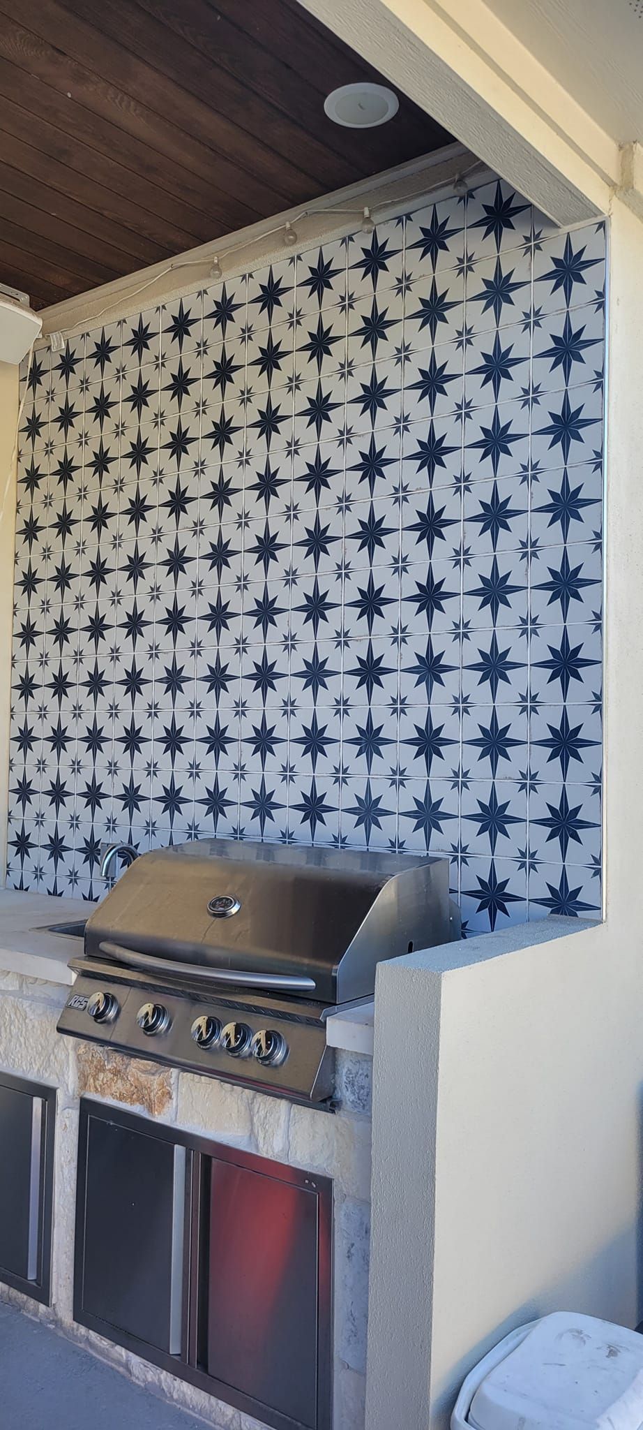 Outdoor kitchen with a built-in grill, stainless steel cabinets, and patterned blue and white tiled backsplash.